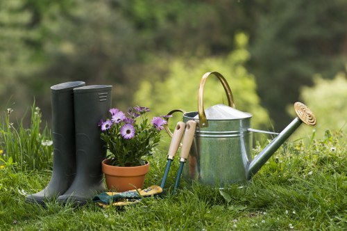 In a lush, green garden setting, a pair of tall, dark green rubber boots is placed on the grass in the foreground. Next to the boots, there is a small terracotta pot filled with purple flowers, adding colour and natural interest. Surrounding these are a variety of gardening tools, including a pair of pruning shears, a hand trowel, and a rake, all arranged neatly on the grass. A silver metal watering can with a curved handle and a long spout is positioned nearby, ready for watering plants and flower beds. The background features blurred greenery, indicating a natural outdoor environment typical of a well-maintained garden space, possibly in the Blackfriars area or nearby London postcodes. The scene appears to be set on a clear, sunny day, with natural daylight illuminating the tools and plants, emphasizing a tidy, organized outdoor workspace suitable for gardening tasks, reflecting professional gardening and landscaping activities often provided by Gardening Services Blackfriars.