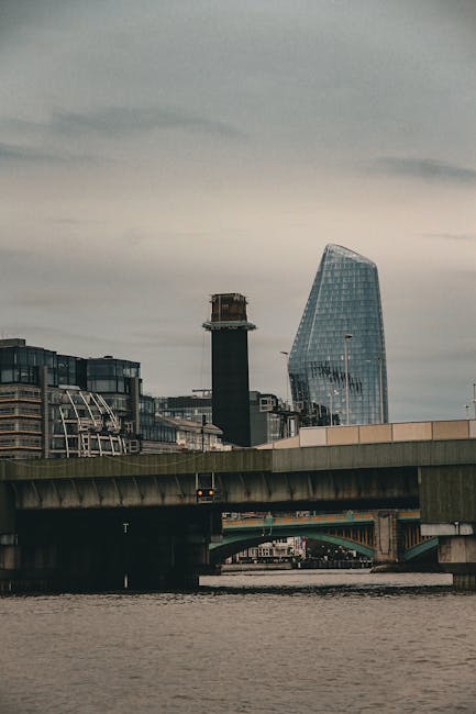 The image depicts an urban riverside scene with a concrete bridge spanning across the water, featuring visible support pillars and a roadway on top. In the background, tall modern buildings are visibl