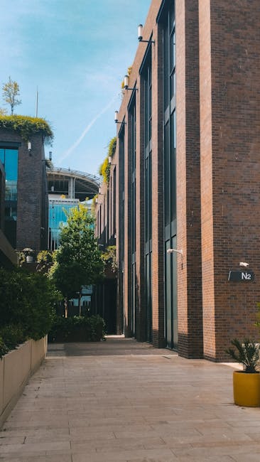 A narrow urban garden pathway runs alongside a modern brick building with large glass windows. The pathway is paved with smooth, light-colored stone tiles, and on the left side, a low concrete wall su