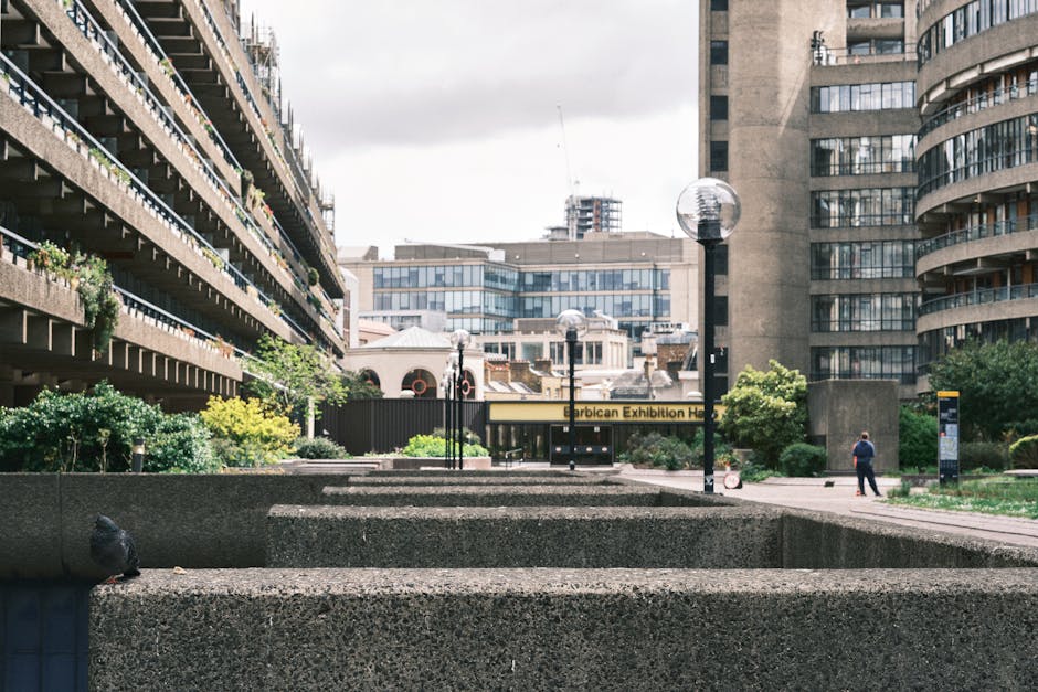 A narrow urban garden pathway runs alongside a modern brick building with large glass windows. The pathway is paved with smooth, light-colored stone tiles, and on the left side, a low concrete wall su