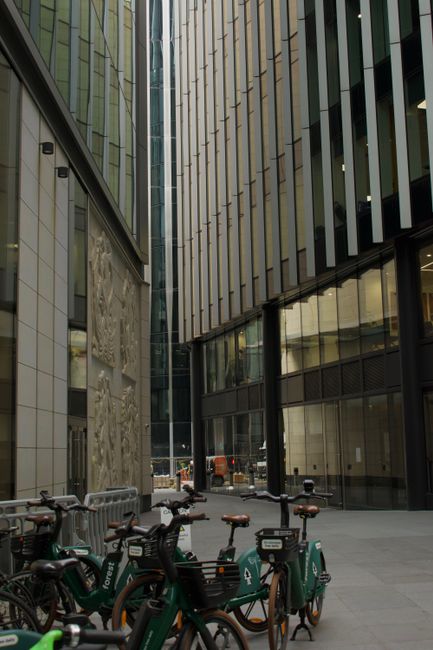 The image depicts a modern urban courtyard situated between tall, contemporary office buildings with glass and metal facades, creating a sleek, reflective environment. Several bicycles, including shar